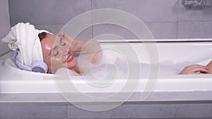 Beautiful young woman takes a bath at home in a modern bathroom. She puts on headphones and listens to her favorite