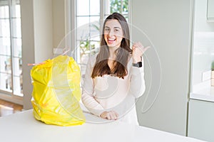 Beautiful young woman taking out the garbage from the rubbish container smiling with happy face looking and pointing to the side