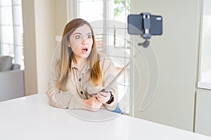 Beautiful young woman taking a picture using selfie stick scared in shock with a surprise face, afraid and excited with fear