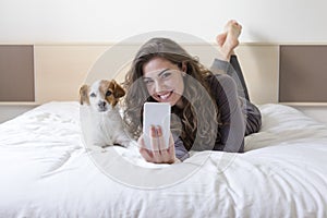 Beautiful young woman taking a selfie with mobile phone on bed with her cute small dog besides. Home, indoors and lifestyle