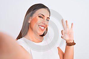 Beautiful young woman taking a selfie photo using smartphone over white background doing ok sign with fingers, excellent symbol