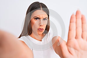 Beautiful young woman taking a selfie photo using smartphone over white background with open hand doing stop sign with serious and