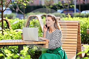 Beautiful young woman talking on mobile phone while using laptop outdoors