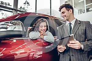 Beautiful young woman is talking to handsome sales manager while choosing a car in dealership