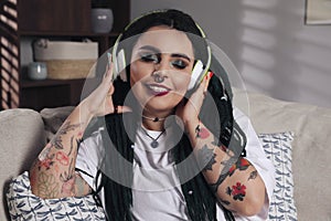 Beautiful young woman with tattoos on body, nose piercing and dreadlocks listening to music at home