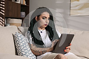 Beautiful young woman with tattoos on body, nose piercing and dreadlocks using tablet at home