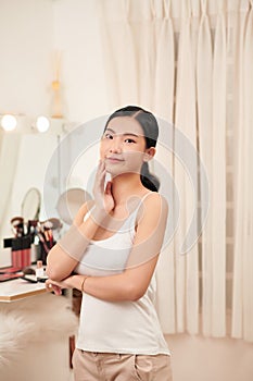 Beautiful young woman is touching her face and smiling while looking at the mirror