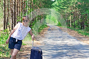 Beautiful young woman tourist with a suitcase on a forest road on a summer sunny day against the backdrop of green trees