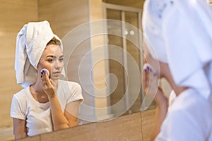 Beautiful young woman is treating her face after shower standing and touching her cheek with a sponge. The woman is holding a