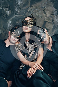 Beautiful young woman and two men in mysterious black mask sitting on floor in studio