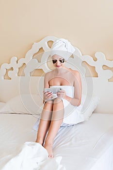 Beautiful young woman with under eye patches and using mobile phone or writing sms massage in bathrobe lying in bed. Happy girl