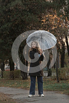 Beautiful young woman under transparent umbrella in autumn park and looks at side. Girl walks on the rain