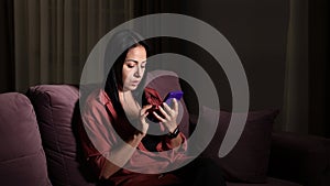 Beautiful young woman use phone sitting on sofa in dark light room at home.
