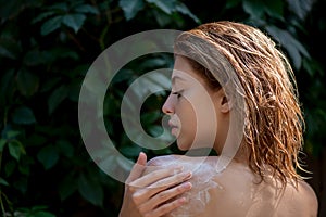 Beautiful young woman uses cream or lotiong on her body front of plant tropical green leaves background