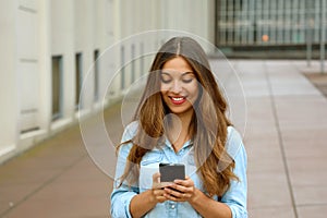 Beautiful young woman is using an app in her smartphone device to send a text message while standing in courtyard of office blocks