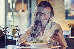 A beautiful young woman is using an application to send an sms message in her smartphone device while eating a salad at the restau