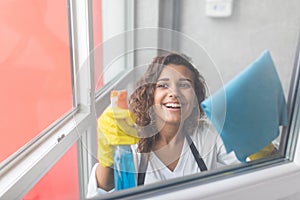 Beautiful young woman is using a duster and a spray, looking at camera and smiling while cleaning windows in the house