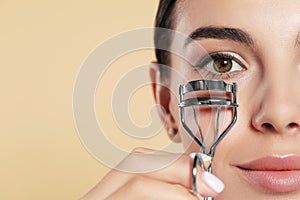 Beautiful young woman using eyelash curler on beige background, closeup. Space for text