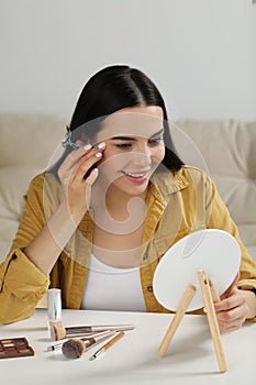 Beautiful young woman using eyelash curler while doing makeup at table indoors