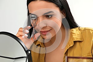 Beautiful young woman using eyelash curler in front of mirror indoors, closeup