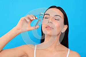 Beautiful young woman using eyelash curler on light blue background