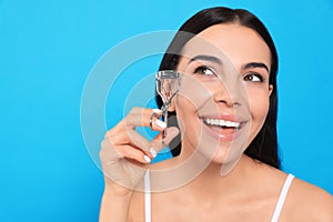 Beautiful young woman using eyelash curler on light blue background, space for text