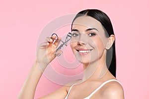 Beautiful young woman using eyelash curler on light pink background