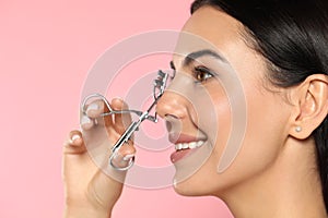 Beautiful young woman using eyelash curler on light pink background, closeup