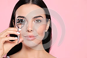 Beautiful young woman using eyelash curler on light pink background, space for text