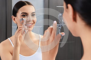 Beautiful young woman using eyelash curler near mirror in bathroom