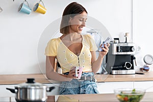 Beautiful young woman using her mobile phone while drinking a cup of coffee in the kitchen at home