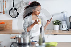 Beautiful young woman using her mobile phone while drinking a cup of coffee in the kitchen at home