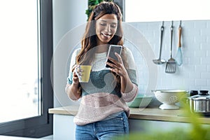 Beautiful young woman using her mobile phone while drinking a cup of coffee in the kitchen at home