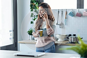 Beautiful young woman using her mobile phone while drinking a cup of coffee in the kitchen at home