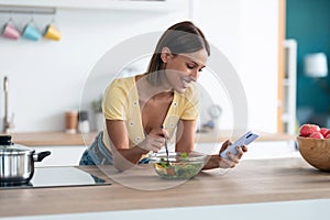 Beautiful young woman using her mobile phone while eating a salad in the kitchen at home