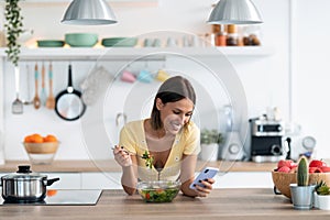 Beautiful young woman using her mobile phone while eating a salad in the kitchen at home