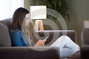 Beautiful young woman using her mobile phone and sitting on sofa at home.