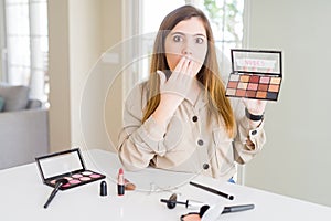 Beautiful young woman using make up cosmetics applying color from palete cover mouth with hand shocked with shame for mistake,