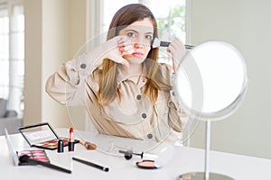 Beautiful young woman using make up cosmetics applying color using brush with angry face, negative sign showing dislike with