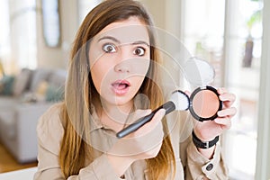 Beautiful young woman using make up cosmetics applying powder scared in shock with a surprise face, afraid and excited with fear