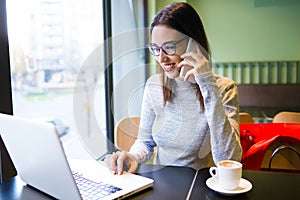 Beautiful young woman using mobile phone while working with her laptop in the coffee shop.