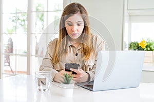 Beautiful young woman using smartphone and computer scared in shock with a surprise face, afraid and excited with fear expression