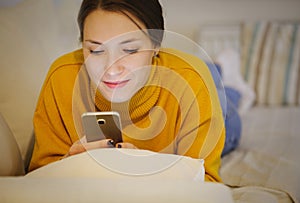 Beautiful young woman using smartphone at home lying on cozy bed