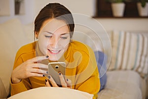 Beautiful young woman using smartphone at home lying on cozy bed