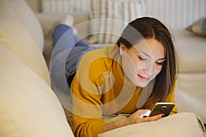 Beautiful young woman using smartphone at home lying on cozy bed