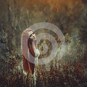 A beautiful young woman with very long red hair as a witch walks through the autumn forest.