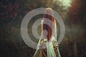 A beautiful young woman with very long red hair as a witch walks through the autumn forest. Back view.