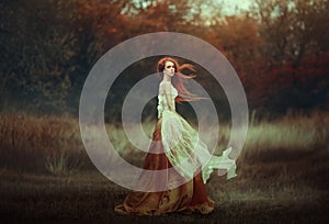 Beautiful young woman with very long red hair in a golden medieval dress walking through the autumn forest. Long red