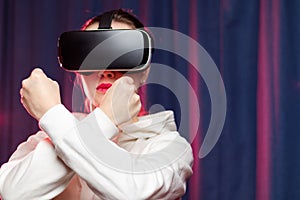 A beautiful young woman in virtual reality glasses and a white hoodie shows stop with her hands on a purple background. Modern tec