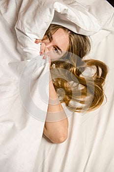 Beautiful young woman wakes up in the morning in white bed in the rays of the sun, she has lush wavy blonde, golden hair